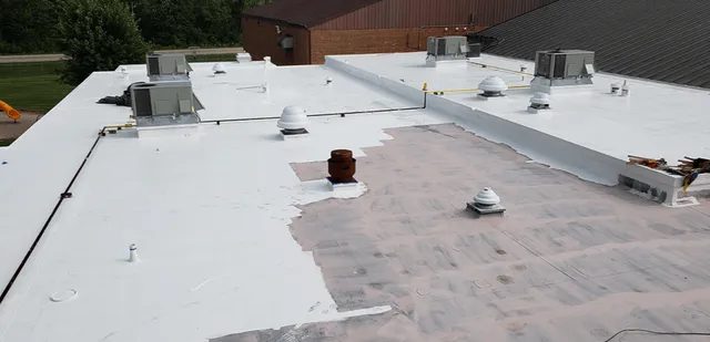 A flat commercial roof partially coated with white sealant, several ventilation units, and HVAC systems; some areas still show exposed underlying material. Trees and another building are visible in the background.