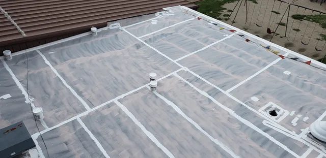 A flat rooftop covered with a white sealant and several ventilation pipes, featuring visible seams and bordered by a playground with swings and a sandy area. Triangular caution flags mark one edge of the roof.