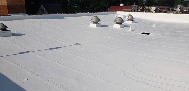 A flat white rooftop with several vent structures and a small dark patch; nearby buildings and trees are visible in the background under a clear sky.