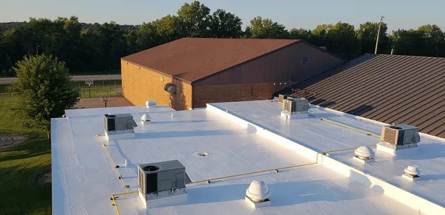 A flat white commercial roof with multiple HVAC units and vents, next to a brown-roofed brick building, surrounded by trees and greenery under a clear sky.