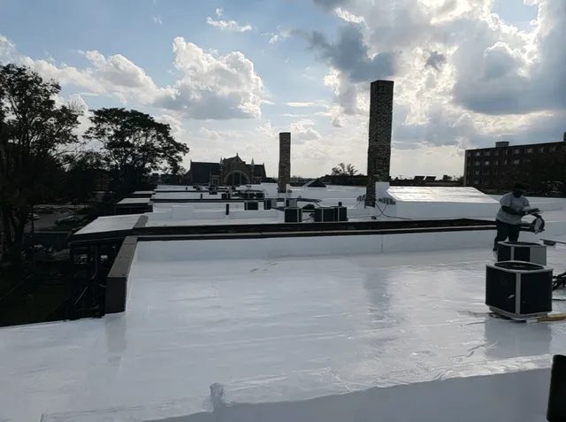 A worker stands on a flat, white rooftop among chimneys and HVAC units. The sky is partly cloudy with sunlight filtering through, and trees and buildings are visible in the background.