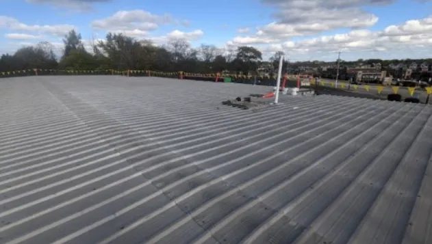 A wide, flat, gray metal rooftop stretches into the distance under a partly cloudy sky, with safety barriers and equipment visible near the center and trees in the background.