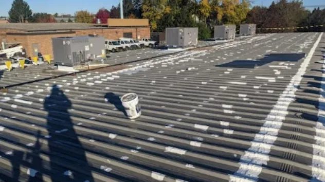 A corrugated metal rooftop with patches of white sealant, several ventilation units, a paint can, and a long shadow of a person. Trees and brick buildings are visible in the background.