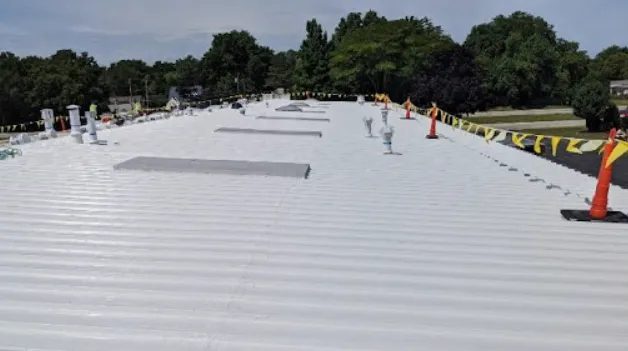 A white metal rooftop with ventilation pipes and orange safety cones, bordered by yellow caution tape. Green trees and a clear sky are visible in the background.