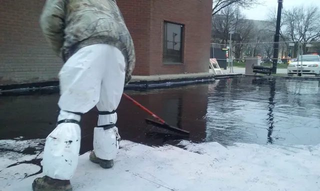 A person wearing white protective clothing uses a broom to spread black sealant over pavement near a brick building, with cars and trees visible in the background.