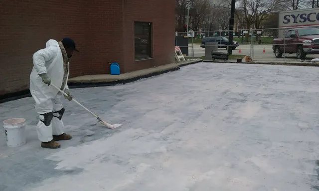A person wearing protective white coveralls and gloves uses a roller to apply a coating to a large, flat concrete surface outside a brick building. Vehicles, a fence, and trees are visible in the background.
