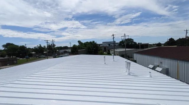 A large white metal roof with several vents sits under a partly cloudy blue sky, surrounded by trees, utility poles, and adjacent industrial buildings.