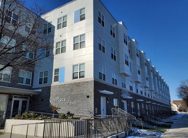 A modern, multi-story apartment building called CityPlace with gray brick and white paneling, large windows, and a ramp leading to the entrance. Snow patches are visible on the ground and steps.