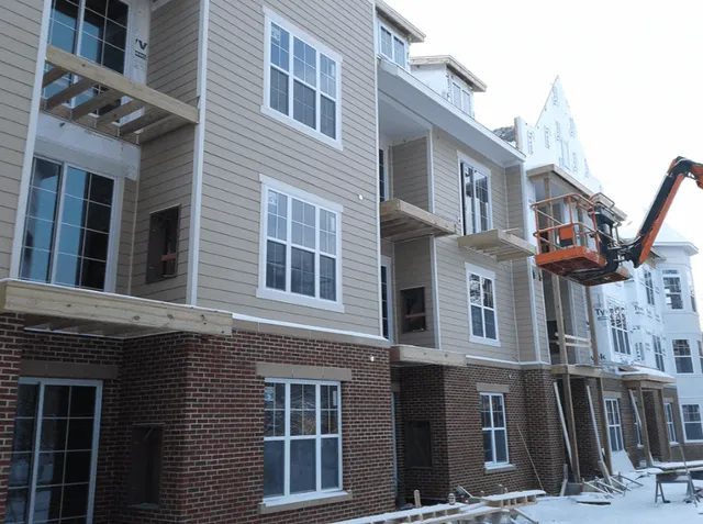 A multi-story apartment building under construction, featuring brick and siding exterior. A boom lift with workers is positioned near the upper floors, and snow covers the ground.
