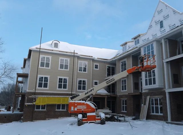 A construction site of a multi-story building in winter; snow covers the roof and ground. An orange JLG lift is raised, with a worker on the platform near upper floors. Building materials are scattered around.