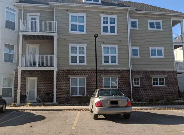 A tan and brick three-story apartment building with balconies and multiple windows. A beige car is parked in front, slightly off-center between two parking spaces. The weather is clear and sunny.