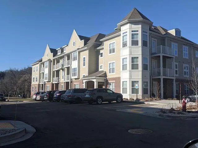 A three-story apartment building with light-colored siding, brick accents, and multiple balconies on a sunny day. Several cars are parked in front, and the sky is clear and blue.